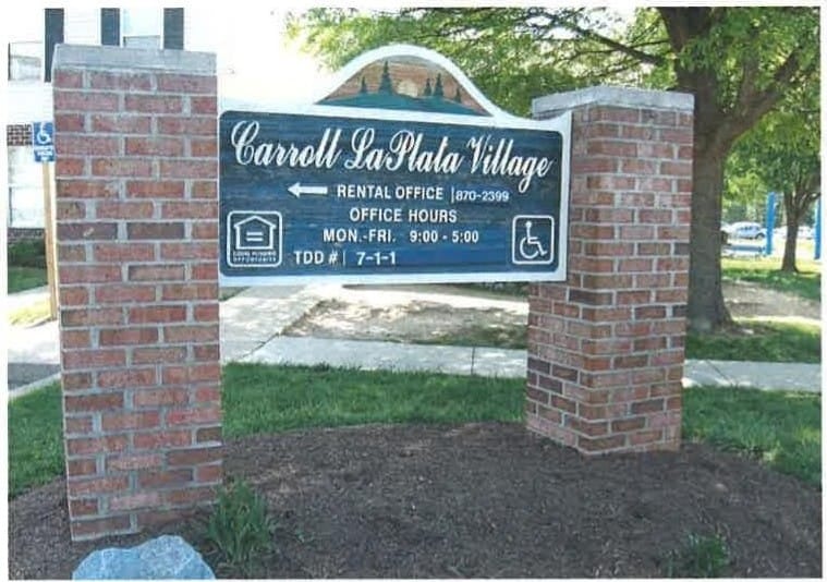 The Carroll LaPlata Village entrance sign on brick pillars, with trees and townhouses behind